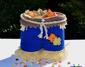 Puede incluir: Un recipiente de ganchillo azul hecho a mano que se asemeja a un pantalón de espantapájaros, lleno de hojas de otoño. El recipiente tiene un borde marrón y está decorado con un botón y lazos de hilo. Flecos de hilo amarillo y caramelos completan el tema otoñal.