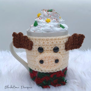 May include: A cream-colored crocheted mug cozy shaped like a moose, with brown antlers and black eyes. The mug is filled with whipped cream, decorated with sprinkles and star-shaped candies. The cozy has a red and green plaid pattern at the bottom.