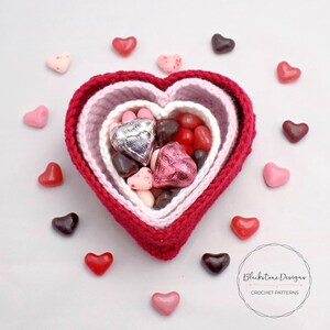 May include: A crocheted heart-shaped basket with three layers in shades of pink and red, filled with heart-shaped candies. The basket is surrounded by scattered heart-shaped candies in pink and red.