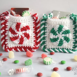 May include: Two crocheted candy cane gift bags with white, red, and green stripes. The bags are filled with small candies and have a loop handle. The bags are sitting on a white surface with scattered candies.