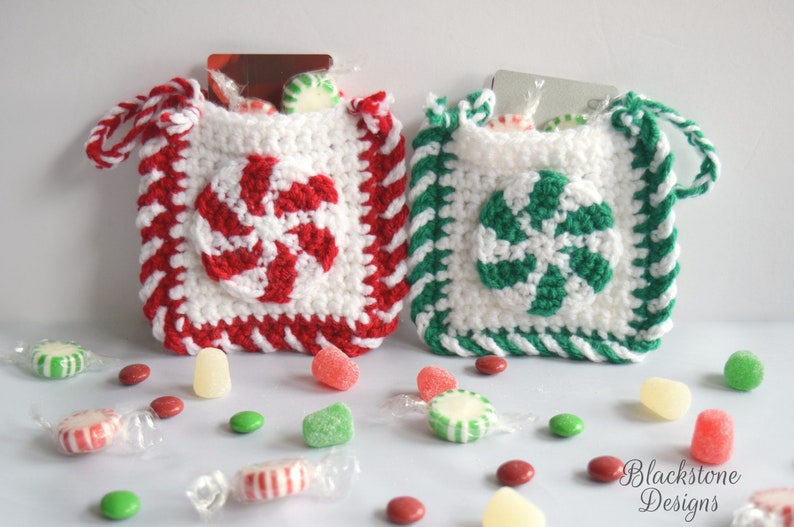 May include: Two crocheted candy cane gift bags with white, red, and green stripes. The bags are filled with small candies and have a loop handle. The bags are sitting on a white surface with scattered candies.