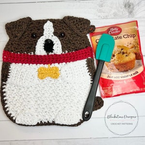 May include: A crochet dog-shaped pot holder in brown, white, and red. The dog has a yellow bone-shaped tag and a black nose. A green spatula and a Betty Crocker Chocolate Chip muffin mix package are next to the pot holder.