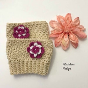 May include: Two beige crochet leg warmers with magenta and white flower accents. The leg warmers are handmade with a ribbed cuff and a textured stitch pattern. A large pink flower is next to the leg warmers. The text "Blackstone Designs" is in the lower right corner.