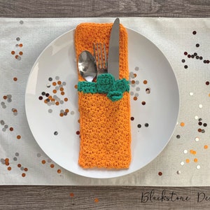 Op de afbeelding: Een witte plaat met bestek in een oranje gehaakte bestekhouder met een groene accent. De plaat staat op een witte placemat met confetti. De tekst "Blackstone Designs" staat onderaan.