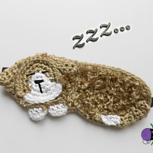 Könnte beinhalten: Eine handgefertigte Schlafmaske aus Häkelarbeit in Form eines schlafenden Hundes. Die Maske ist hauptsächlich hellbraun mit weißen Akzenten auf Gesicht und Pfoten. Das Wort "ZZZ..." steht in silbernen Buchstaben über der Maske. Die Maske ist zum Abdunkeln konzipiert.