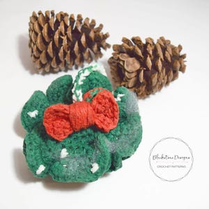 May include: A handmade green crochet wreath with a red bow and white accents. Two brown pinecones are in the background. The wreath is decorated with white glitter. The text "Blackstone Designs CROCHET PATTERNS" is in a circle.
