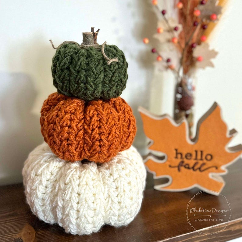 May include: A stack of three crocheted pumpkins in green, orange, and white. The pumpkins are stacked on a wooden surface with a wooden leaf sign that says "hello fall".