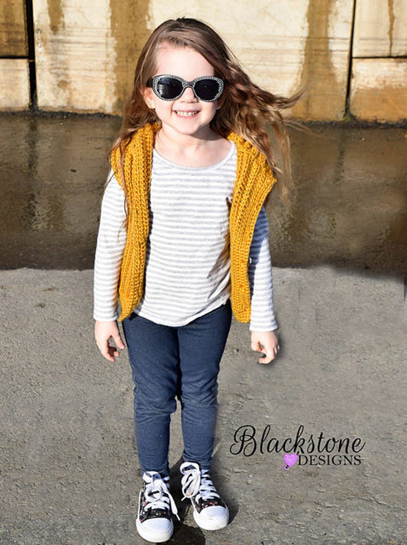 May include: A young person wearing a yellow crocheted vest over a striped shirt, blue jeans, and black and white sneakers. The person is wearing black sunglasses and has long brown hair.