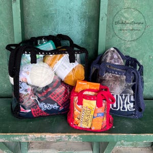 May include: Three clear plastic tote bags with black, blue, and red handles. The bags are filled with yarn, fabric, and snacks. The bags are sitting on a green bench. The text "Blackstone Designs Crochet Patterns" is visible in the top right corner of the image.