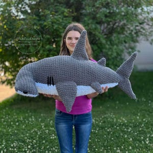 May include: A crocheted hammerhead shark plush toy in gray and white, held by a person wearing a pink shirt and blue jeans. The plush toy has a large head with a flat, hammer-shaped snout and a white underbelly.