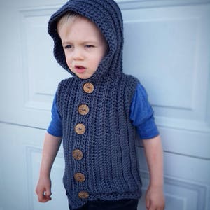 May include: A gray hooded vest with wooden buttons. The vest is crocheted and has a ribbed texture. The vest is being worn by a child.