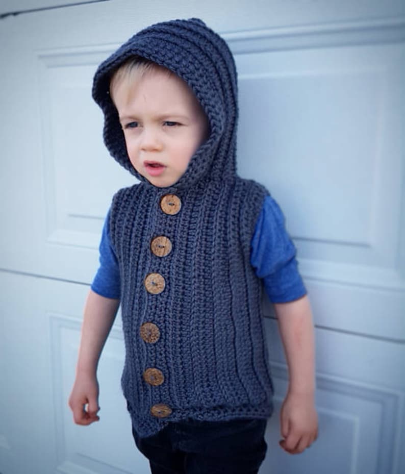 May include: A gray hooded vest with wooden buttons. The vest is crocheted and has a ribbed texture. The vest is being worn by a child.