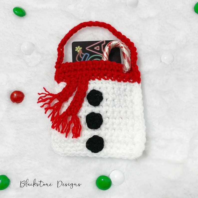 May include: White crocheted snowman gift bag with a red scarf and a candy cane inside. The bag has black buttons for eyes and a nose.