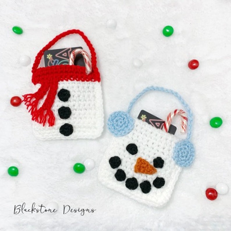 May include: Two crocheted snowman gift bags with white yarn, black buttons, and orange noses. One bag has a red scarf and blue pom-poms, the other has blue pom-poms. Both bags have a gift card and candy cane inside.