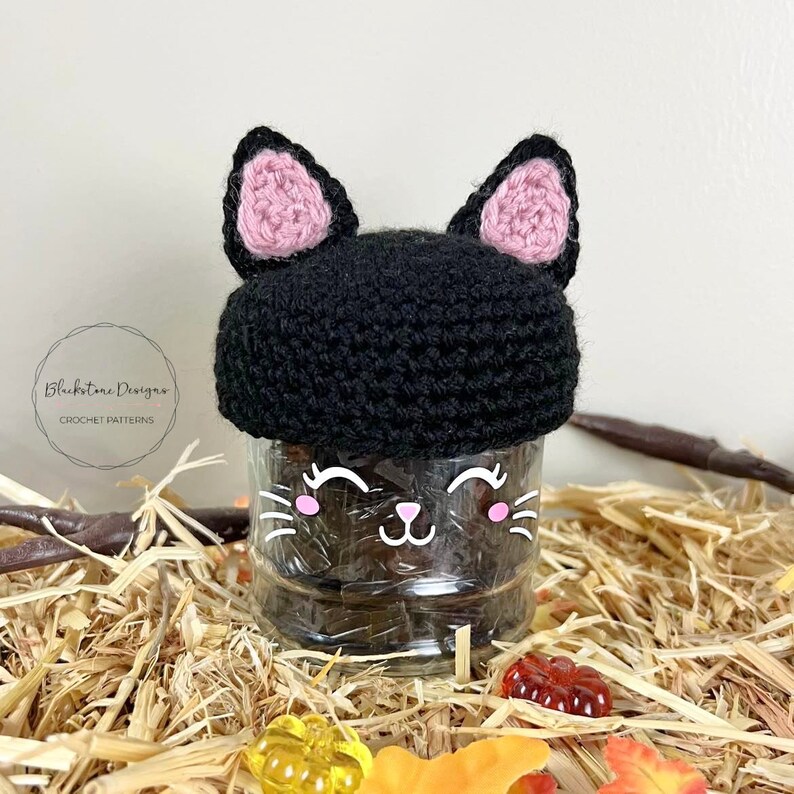 K&ouml;nnte beinhalten: Eine schwarze geh&auml;kelte Katzenm&uuml;tze mit rosa Ohren sitzt auf einem durchsichtigen Glasgef&auml;&szlig; mit einem niedlichen Katzenkopf darauf. Das Gef&auml;&szlig; ist von Stroh und Herbstlaub umgeben.