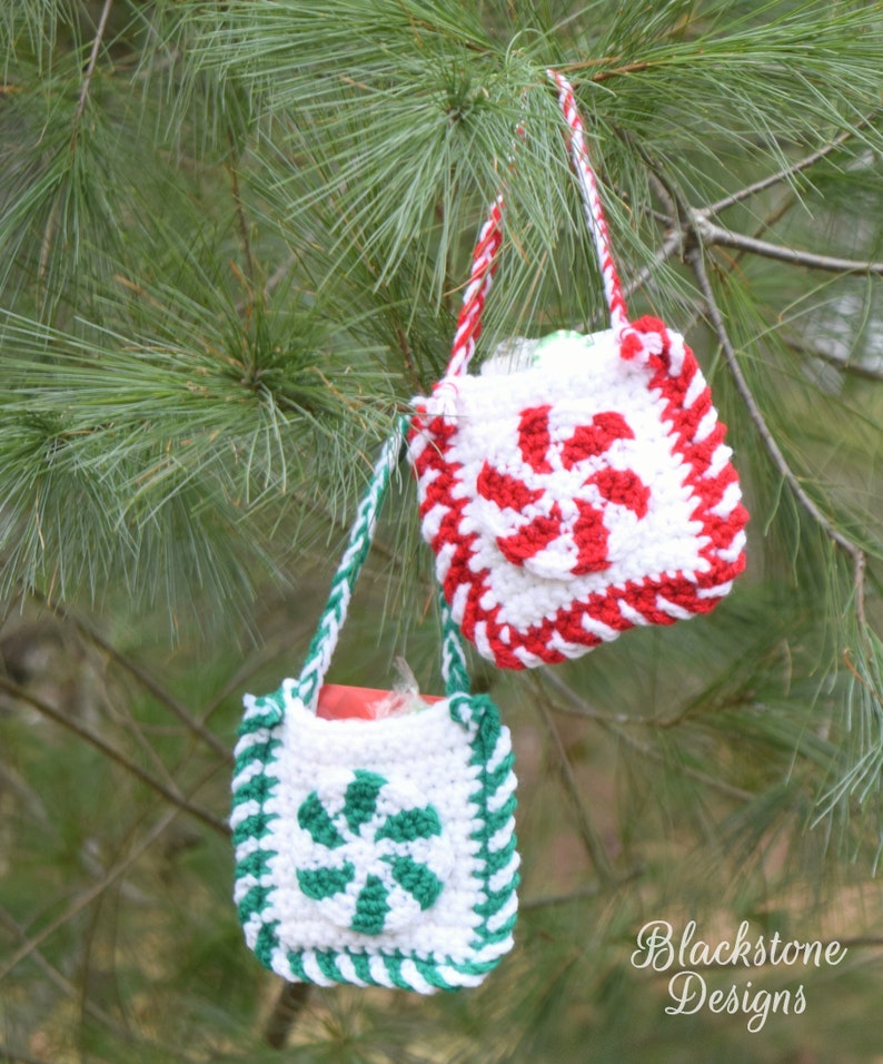 May include: Two crocheted candy cane gift bags hanging from a pine tree branch. The bags are white with red and green stripes and a candy cane design in the center. The bags are perfect for small gifts or treats.
