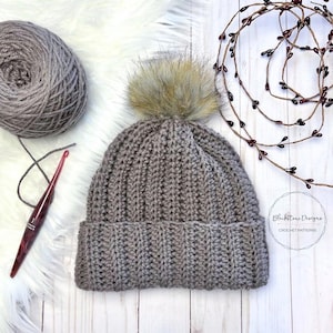 May include: A brown crocheted beanie with a faux fur pom pom on top. The beanie is on a white wooden surface with a ball of brown yarn and a crochet hook.