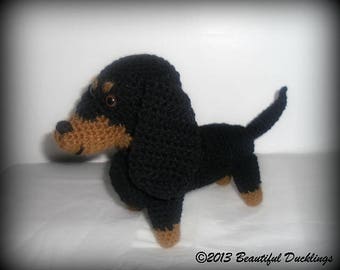 May include: A handmade crochet dachshund dog in black and tan. The dog has a long body, floppy ears, and a small tail. The dog is standing on a white surface.