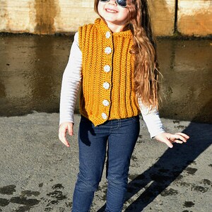 May include: A young girl wearing a yellow crocheted vest with white buttons, blue jeans, and black sneakers. She is wearing sunglasses and has long brown hair.