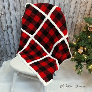 Puede incluir: Manta de crochet de cuadros rojos y negros con borde blanco. La manta está doblada y drapeada sobre una silla blanca. La manta está hecha con un hilo suave y esponjoso.