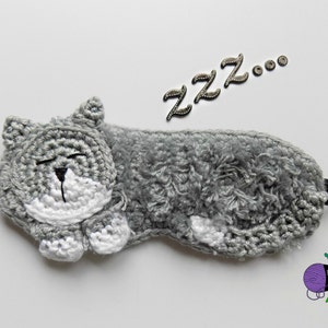 May include: A handmade gray crochet sleep mask shaped like a sleeping cat, with white accents and a black elastic band. The word "ZZZ..." is above the mask. A sleep accessory.