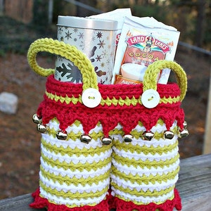 Puede incluir: Una bolsa de regalo de crochet con rayas rojas y verdes, ribete blanco y cascabeles. La bolsa está llena de una lata plateada y dos paquetes de Land O'Lakes Cocoa Classic.