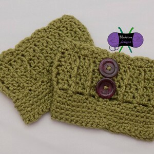 May include: Two olive green crochet wrist cuffs with a button closure. Each cuff is made of a textured stitch and features two dark purple buttons. The cuffs are handmade and suitable for cold weather.