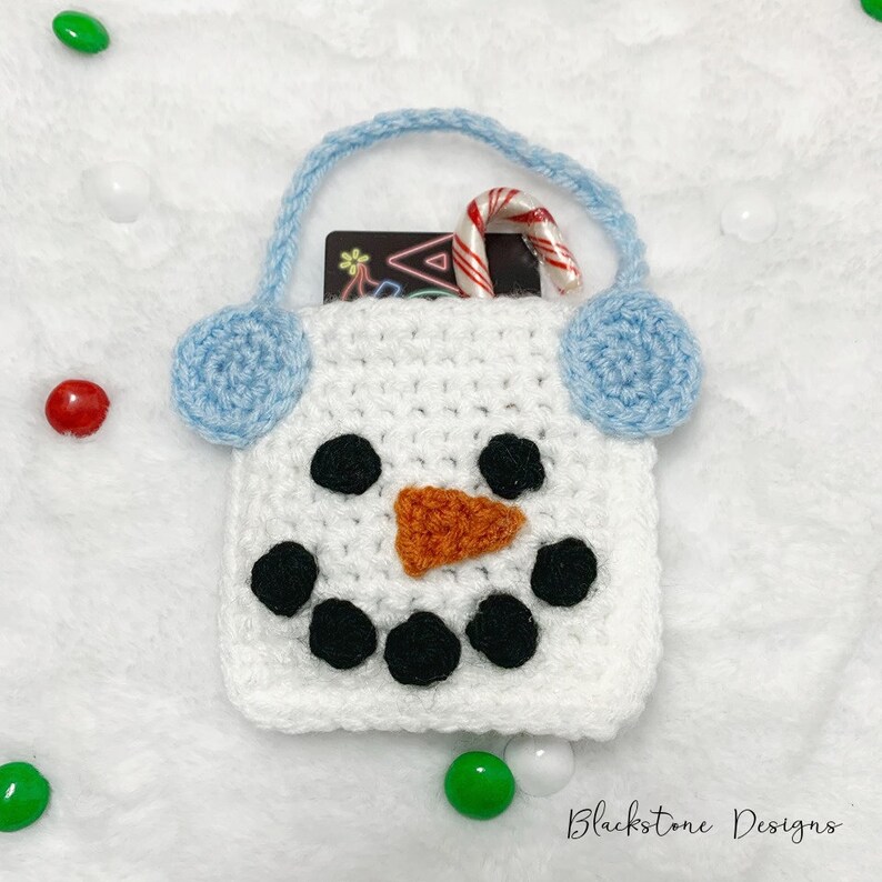 May include: White crocheted snowman gift bag with blue earmuffs, a candy cane, and a gift card inside. The bag has black eyes, a black mouth, and an orange nose.