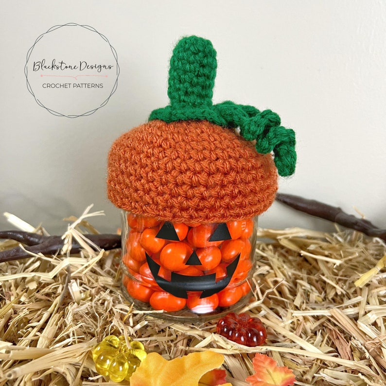 K&ouml;nnte beinhalten: Ein geh&auml;kelter orangefarbener K&uuml;rbis-Hut mit einem gr&uuml;nen Stiel und Blatt sitzt auf einem Glasgef&auml;&szlig;, das mit orangefarbenen S&uuml;&szlig;igkeiten gef&uuml;llt ist. Das Gef&auml;&szlig; hat ein schwarzes K&uuml;rbisgesicht darauf gezeichnet. Das Gef&auml;&szlig; steht auf einem Strohboden.
