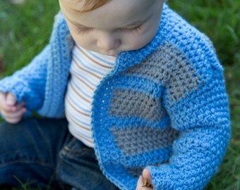 May include: A baby wearing a blue and gray crochet cardigan over a striped shirt and blue jeans. The cardigan has a textured stitch pattern and a contrasting gray design on the front. The child is sitting on green grass.