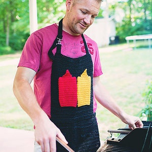 Op de afbeelding: Een man draagt een zwarte en rode gehaakte schort met een ontwerp van twee ketchup- en mosterdflessen. De schort heeft verstelbare banden en een zak. Hij staat voor een zwarte grill.