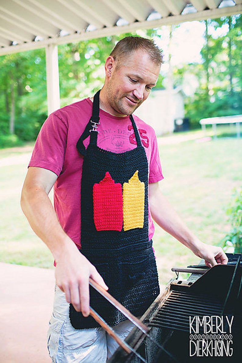 Op de afbeelding: Een man draagt een zwarte en rode gehaakte schort met een ontwerp van twee ketchup- en mosterdflessen. De schort heeft verstelbare banden en een zak. Hij staat voor een zwarte grill.