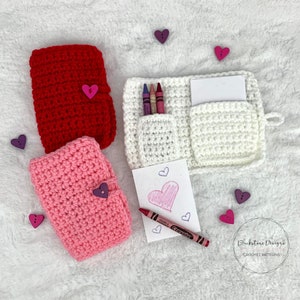 May include: Three crocheted pouches in red, pink, and white with heart-shaped buttons. The white pouch has a pocket for crayons and a small notepad. The pouches are on a white fuzzy surface with hearts scattered around.