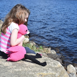 May include: A young girl sits on a rock by a lake, holding a pink crocheted flamingo plush toy. The flamingo has black eyes and a black beak.