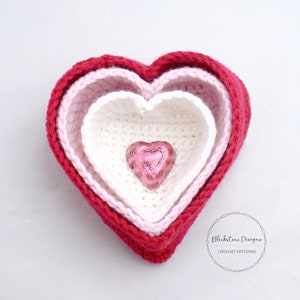 May include: Three crocheted heart-shaped bowls, nested inside each other, in shades of red, pink, and white. A small heart-shaped chocolate candy is inside the smallest bowl.
