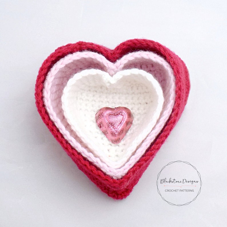 May include: Three crocheted heart-shaped bowls, nested inside each other, in shades of red, pink, and white. A small heart-shaped chocolate candy is inside the smallest bowl.