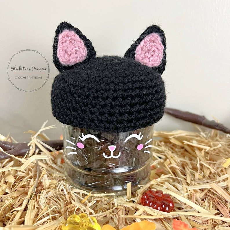 K&ouml;nnte beinhalten: Eine schwarze geh&auml;kelte Katzenm&uuml;tze mit rosa Ohren sitzt auf einem durchsichtigen Glasgef&auml;&szlig; mit einem niedlichen Comic-Katzenkopf darauf. Das Gef&auml;&szlig; ist von Stroh und bunten S&uuml;&szlig;igkeiten umgeben.