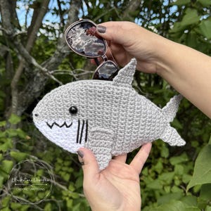 May include: A crocheted shark-shaped pouch in light gray, with a white mouth and black accents. The pouch has a zipper closure and is held in a person's hand. Sunglasses are placed on top of the pouch. The background is blurred green foliage.