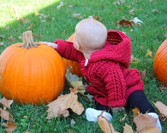 May include: A baby in a red hooded sweater and black pants sits near large orange pumpkins on a grassy lawn. The sweater has a white button closure. Autumn leaves are scattered around the pumpkins.
