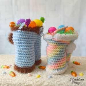 May include: Two crocheted Easter baskets, one blue and one rainbow, filled with colorful plastic eggs and Easter grass. The baskets are standing on a white fluffy surface.
