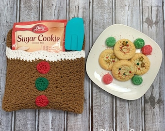 May include: A crocheted gingerbread house-shaped bag with red and green buttons, filled with a box of Betty Crocker Sugar Cookie mix and a blue spatula. A white plate with sugar cookies and red and green candies sits on a woodgrain surface.