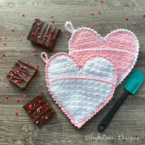 May include: Two heart-shaped crochet potholders in white and pink, with a white trim. Brownies with chocolate frosting and sprinkles are scattered around the potholders. A teal spatula with a black handle is also present. The text "Blackstone Designs" is at the bottom.