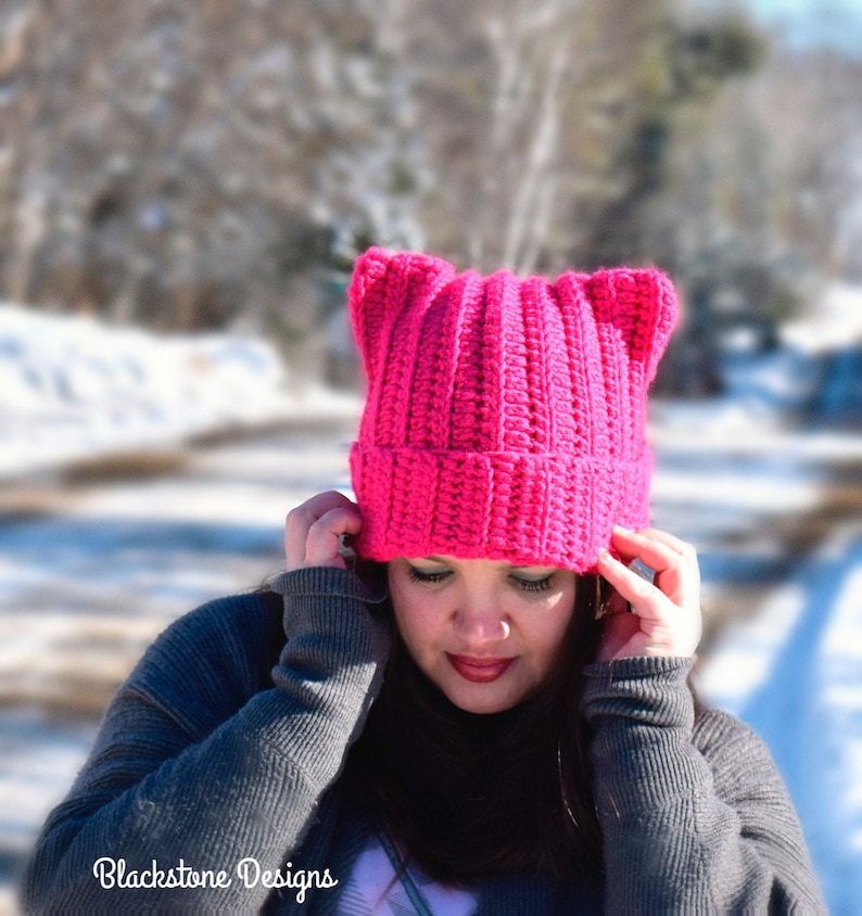 May include: A pink crocheted hat with cat ears. The hat is worn by a person with dark hair and a gray sweater.