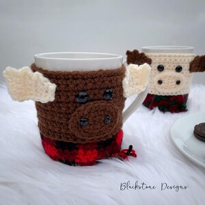 May include: Two white ceramic mugs with crocheted moose cozies. The brown cozy has cream-colored antlers and a red and black plaid bottom. The second mug has a cream-colored cozy with brown antlers and a green and red plaid bottom. The mugs are on a white, fluffy surface.