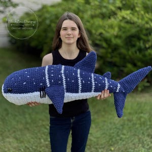 Könnte beinhalten: Ein großer, handgefertigter Walhai-Plüsch aus Häkelarbeit. Der Plüsch ist überwiegend dunkelblau mit weißen Streifen und Punkten und einem weißen Bauch. Der Text "Blackstone Designs CROCHET PATTERNS" ist sichtbar.