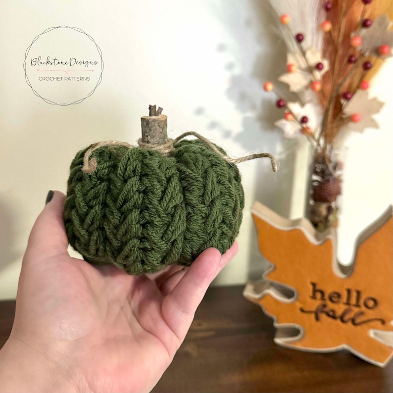 May include: A green crocheted pumpkin with a twig stem and twine accents. The pumpkin is being held in a hand, with a wooden "hello fall" sign in the background.