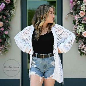 Op de afbeelding: Een witte, open geweven gehaakte cardigan met lange mouwen. De cardigan wordt gedragen over een zwarte top en een spijkerbroek. Een zwarte riem met een gouden gesp is zichtbaar. De tekst "Blackstone Designs Crochet Patterns" is inbegrepen.