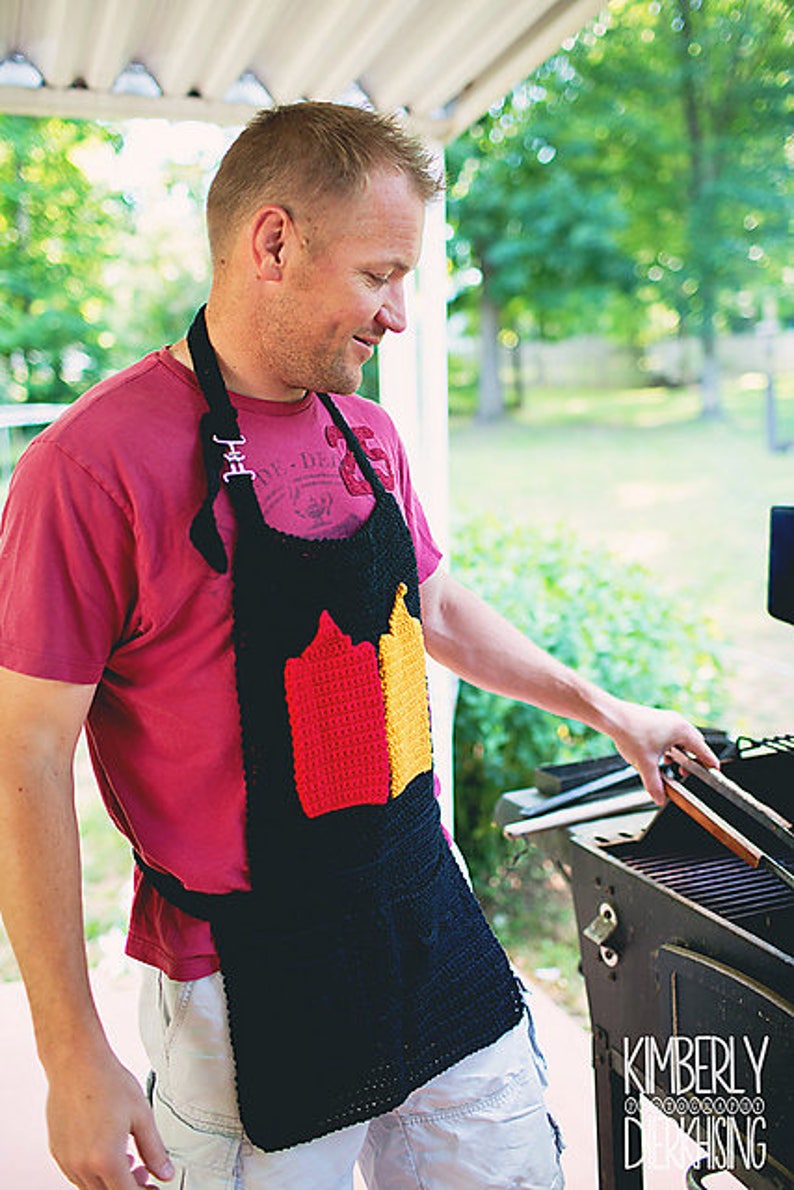 Op de afbeelding: Een persoon draagt een rood shirt met een zwart en rood gebreid schort met een ontwerp dat lijkt op de Australische vlag. Het schort heeft een zwarte band en een zwarte zak. De persoon staat voor een zwarte grill.