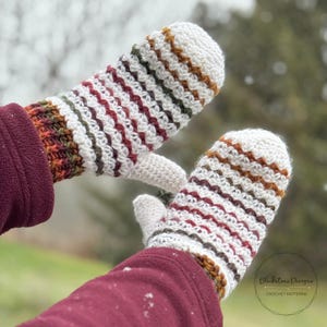 Könnte beinhalten: Ein Paar handgefertigte Häkelhandschuhe. Die Handschuhe sind weiß mit horizontalen Streifen in Rot, Braun, Grün und Orange. Die Bündchen sind aus dunklerem, strukturiertem Garn. Warme und stilvolle Fäustlinge.