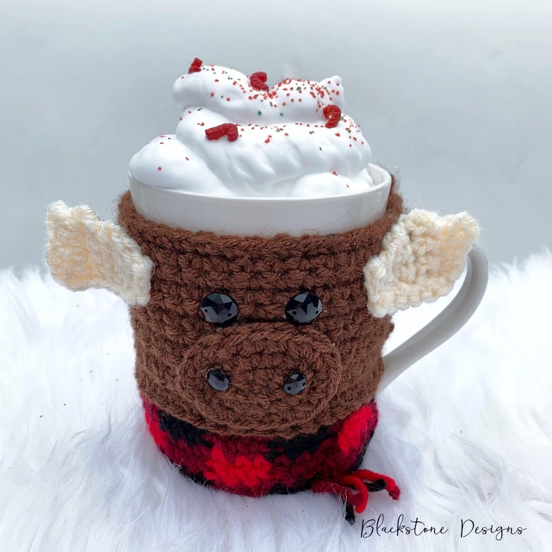 May include: A white mug with a brown crocheted cozy shaped like a bull, complete with black button eyes and cream-colored wings. The mug is filled with whipped cream and red and green sprinkles. The cozy has a red and black plaid pattern.
